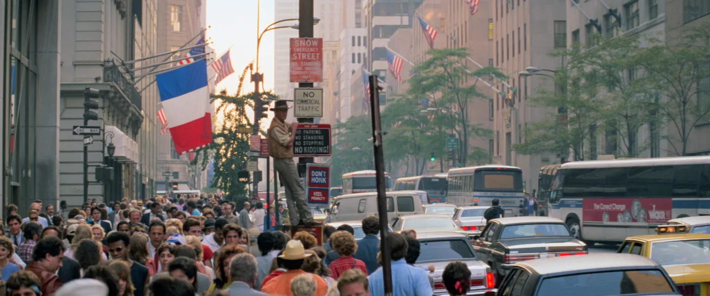 Eine belebte Straßenszene in einer Großstadt mit vielen Menschen auf dem Bürgersteig und mehreren Fahrzeugen auf der Straße. Im Vordergrund ist eine Person auf einem Schildpfosten stehend zu sehen, die eine französische Flagge hält. Im Hintergrund sind hohe Gebäude und Bäume zu erkennen. Verkehrsschilder mit Hinweisen wie "No Standing Anytime" und "No Stopping Anytime" sind sichtbar.  [erstellt mit KI]