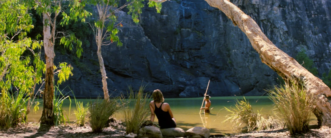 Eine Person sitzt auf einem Felsen am Ufer eines ruhigen, grünen Gewässers, umgeben von dichter Vegetation mit Bäumen und hohen Gräsern. Im Wasser steht eine weitere Person, die eine lange Stange hält. Im Hintergrund erhebt sich eine steile, felsige Klippe. [erstellt mit KI]