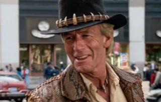 Person mit schwarzem Hut, der mit Zähnen oder Klauen verziert ist, trägt eine braune Jacke mit Schlangenledermuster und ein beiges Hemd. Im Hintergrund sind ein Gebäude mit großen Fenstern, ein rotes Auto und weitere Fahrzeuge zu sehen. Die Szene spielt sich im Freien ab. [erstellt mit KI]