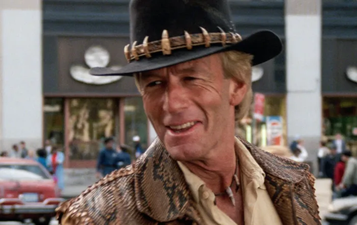 Person mit schwarzem Hut, der mit Zähnen oder Klauen verziert ist, trägt eine braune Jacke mit Schlangenledermuster und ein beiges Hemd. Im Hintergrund sind ein Gebäude mit großen Fenstern, ein rotes Auto und weitere Fahrzeuge zu sehen. Die Szene spielt sich im Freien ab. [erstellt mit KI]