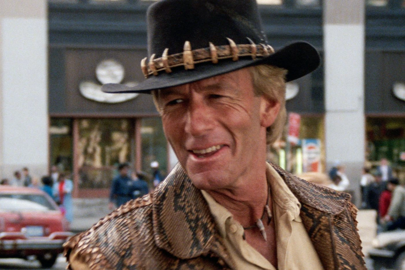 Person mit schwarzem Hut, der mit Zähnen oder Klauen verziert ist, trägt eine braune Jacke mit Schlangenledermuster und ein beiges Hemd. Im Hintergrund sind ein Gebäude mit großen Fenstern, ein rotes Auto und weitere Fahrzeuge zu sehen. Die Szene spielt sich im Freien ab. [erstellt mit KI]