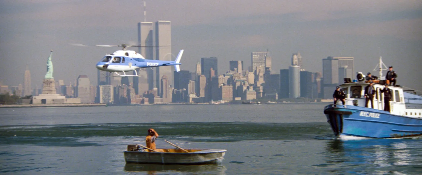 Im Vordergrund rudert eine Person in einem kleinen Ruderboot auf dem Wasser. Rechts fährt ein größeres Polizeiboot mit mehreren Personen an Bord. Links ist die Freiheitsstatue zu sehen. Im Hintergrund fliegt ein Hubschrauber mit der Aufschrift 'POLICE' vor der Skyline von New York, darunter die Zwillingstürme des World Trade Centers. [erstellt mit KI]