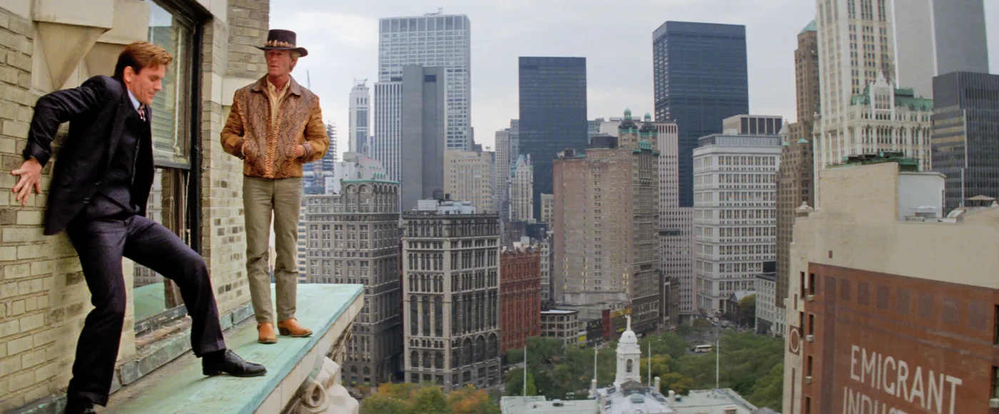 Zwei Personen stehen auf einem Sims eines hohen Gebäudes in einer Großstadt. Eine Person trägt einen dunklen Anzug mit Krawatte, die andere eine gemusterte Jacke und einen Hut. Im Hintergrund sind zahlreiche Hochhäuser und Wolkenkratzer zu sehen. [erstellt mit KI]