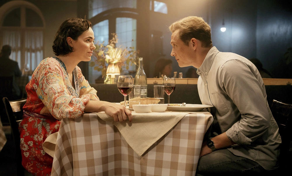 Zwei Personen sitzen sich an einem mit einem karierten Tischtuch bedeckten Tisch gegenüber. Auf dem Tisch stehen zwei Gläser mit Rotwein, eine Wasserflasche und ein Brotkorb. Im Hintergrund sind Fenster mit Licht und eine Vase mit getrockneten Blumen zu sehen. Die Szene wirkt gemütlich und warm beleuchtet. [erstellt mit KI]