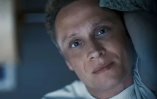 Ein Mann mit lockigem, hellbraunem Haar trägt ein weißes T-Shirt und liegt auf der Seite in einem dunklen Raum. Im Hintergrund ist eine Wand mit einem Fenster oder einer hellen Fläche zu sehen. [erstellt mit KI]