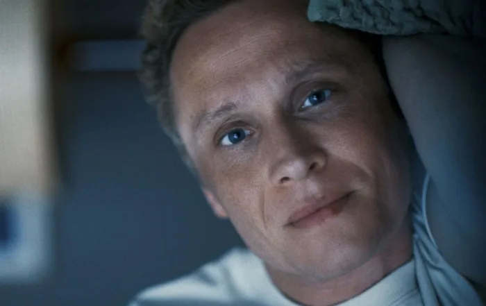 Ein Mann mit lockigem, hellbraunem Haar trägt ein weißes T-Shirt und liegt auf der Seite in einem dunklen Raum. Im Hintergrund ist eine Wand mit einem Fenster oder einer hellen Fläche zu sehen. [erstellt mit KI]