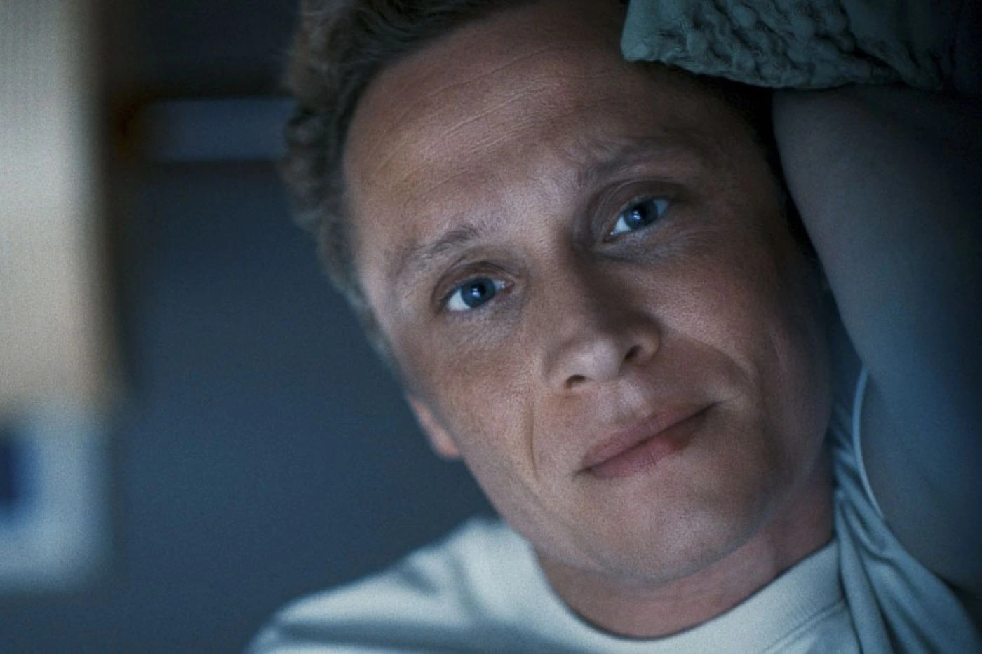 Ein Mann mit lockigem, hellbraunem Haar trägt ein weißes T-Shirt und liegt auf der Seite in einem dunklen Raum. Im Hintergrund ist eine Wand mit einem Fenster oder einer hellen Fläche zu sehen. [erstellt mit KI]