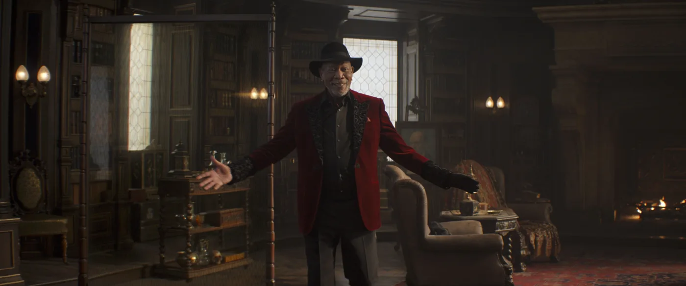 Eine Person mit schwarzem Hut und rotem Sakko steht mit ausgebreiteten Armen in einem elegant eingerichteten Raum. Im Hintergrund sind Bücherregale, ein Sessel, ein Tisch mit Kerzen und weitere klassische Möbelstücke zu sehen. [erstellt mit KI]