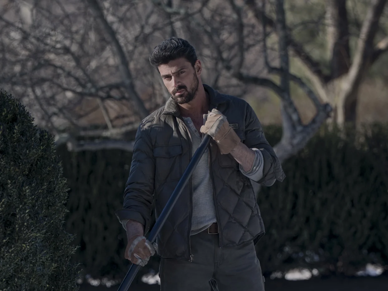 Eine Person steht im Freien und hält mit beiden Händen einen langen Stab oder Rohr. Die Person trägt eine gesteppte, dunkle Jacke, ein helles Oberteil und Arbeitshandschuhe. Im Hintergrund sind kahle Äste und eine dichte Hecke zu sehen. Die Szene wirkt winterlich oder frühjährlich. [erstellt mit KI]