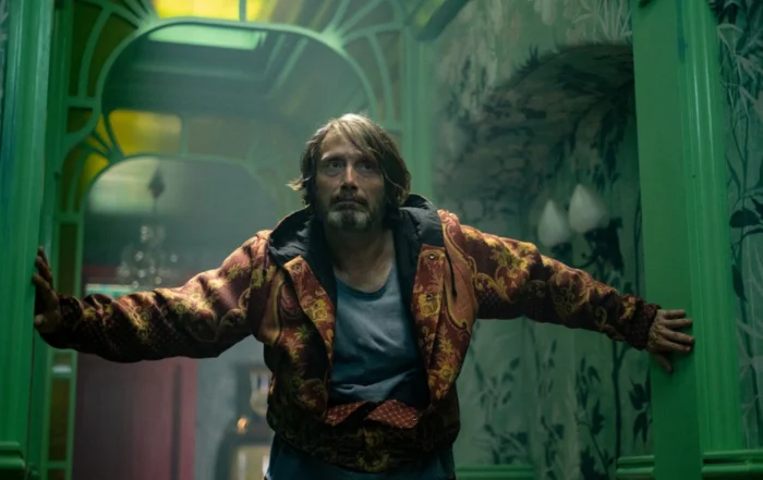 Eine Person mit ausgestreckten Armen steht in einem Raum mit grünen Türrahmen und floraler Tapete. Die Person trägt eine gemusterte Jacke in Rot- und Gelbtönen über einem grauen T-Shirt. Im Hintergrund sind Lampen und ein gelb beleuchtetes Oberlicht zu sehen. [erstellt mit KI]