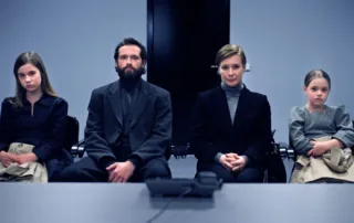 Vier Personen sitzen nebeneinander auf Stühlen vor einem schwarzen Hintergrund. Sie tragen dunkle, formelle Kleidung, darunter Anzüge und Pullover. Ihre Hände sind vor ihnen gefaltet oder liegen auf den Knien. Im Vordergrund liegt ein Tisch, auf dem ein Mikrofon steht. [erstellt mit KI]