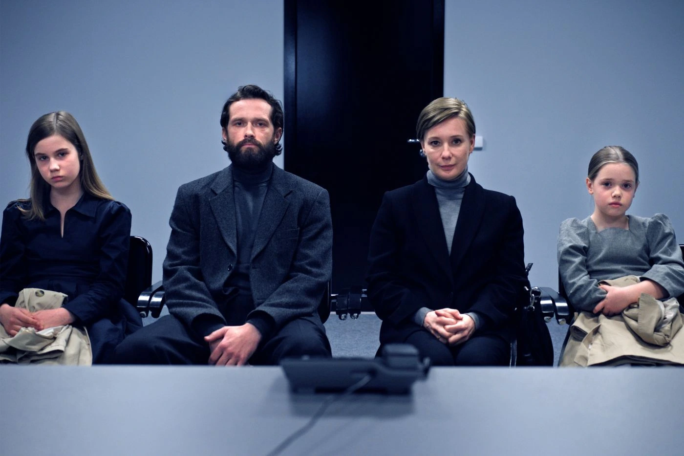 Vier Personen sitzen nebeneinander auf Stühlen vor einem schwarzen Hintergrund. Sie tragen dunkle, formelle Kleidung, darunter Anzüge und Pullover. Ihre Hände sind vor ihnen gefaltet oder liegen auf den Knien. Im Vordergrund liegt ein Tisch, auf dem ein Mikrofon steht. [erstellt mit KI]