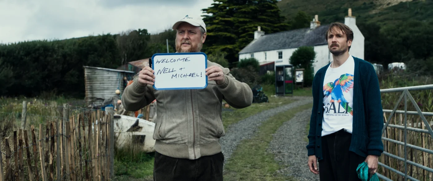 Zwei Männer stehen auf einem schmalen Weg vor ländlicher Kulisse, während einer von ihnen ein handgeschriebenes Schild mit der Aufschrift „Welcome Nell + Michael!“ in die Kamera hält.
