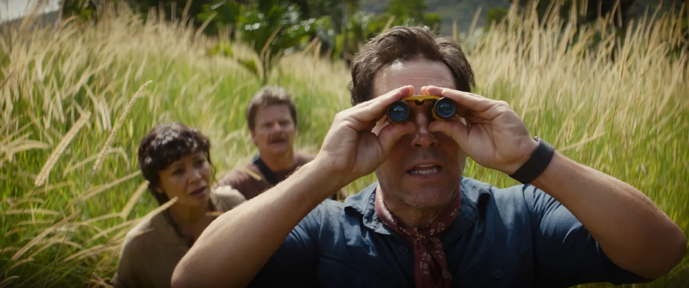 Drei Personen stehen in einem Feld mit hohem, gelblich-grünem Gras. Die vorderste Person trägt ein dunkelblaues Hemd und ein rotes Halstuch und hält sich die Hände an die Ohren. Die beiden Personen im Hintergrund tragen dunkle Kleidung. [erstellt mit KI]