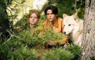 Ein Junge, ein Mädchen und ein schneeweißer Wolf hocken hinter einem Gestrüpp im Wald