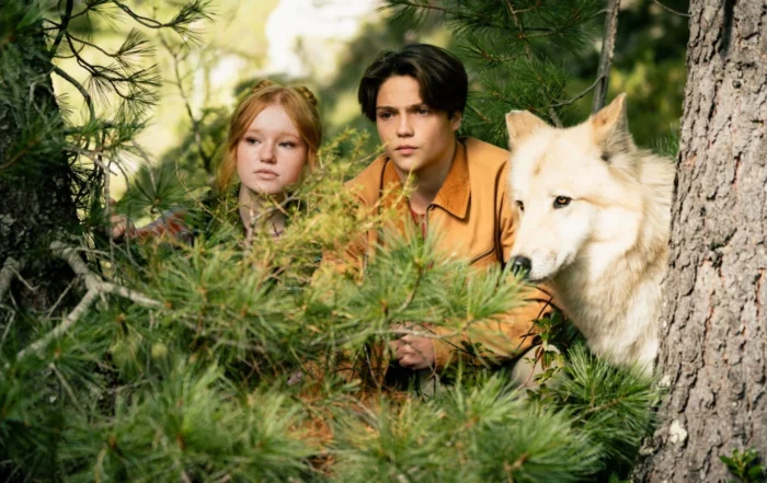 Ein Junge, ein Mädchen und ein schneeweißer Wolf hocken hinter einem Gestrüpp im Wald