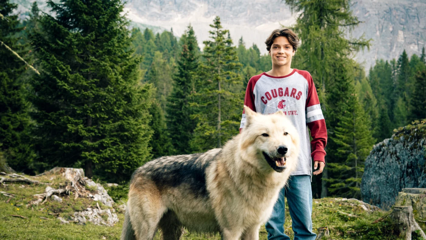 ein Junge steht auf einer Lichtung im Wald, vor ihm eine große Schneewölfin, beide schauen in Richtung Kamera
