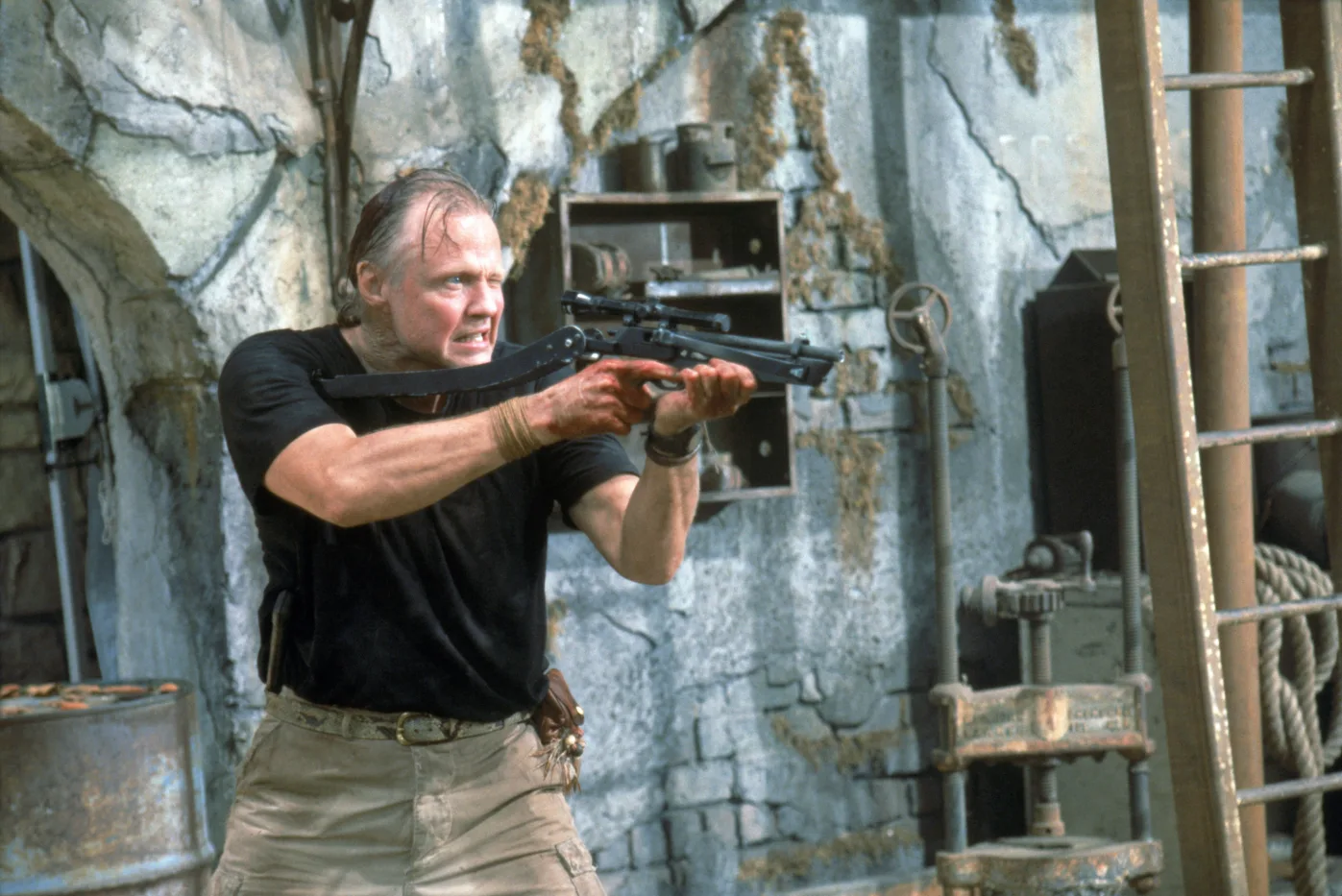 Ein Mann in schwarzem T-Shirt und beiger Hose hält eine Schusswaffe mit Zielfernrohr in beiden Händen und zielt nach vorne. Er steht vor einer verwitterten, grauen Betonwand mit sichtbaren Rissen und Rostflecken. Im Hintergrund sind ein Metallregal mit verschiedenen Gegenständen, ein altes Rohrsystem und ein großer Metallzylinder zu sehen. [erstellt mit KI]