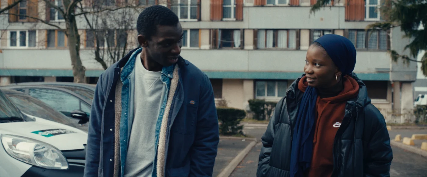 Zwei junge Menschen gehen nebeneinander durch eine Wohnsiedlung und unterhalten sich, im Hintergrund sind geparkte Autos und ein Mehrfamilienhaus zu sehen.