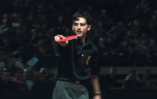 Eine Person in dunkler Kleidung steht in einer Halle vor einem unscharfen Publikum und hält einen roten Tischtennisschläger mit ausgestrecktem Arm nach vorne. Auf dem Hemd ist ein auffälliges Abzeichen zu sehen.
