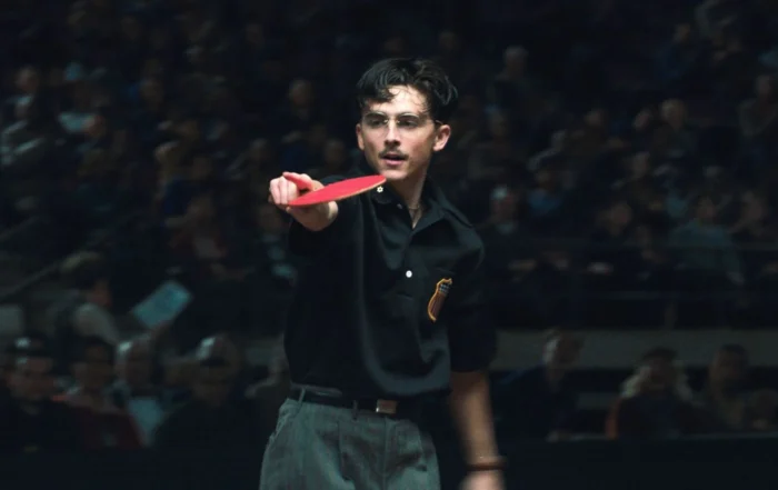 Eine Person in dunkler Kleidung steht in einer Halle vor einem unscharfen Publikum und hält einen roten Tischtennisschläger mit ausgestrecktem Arm nach vorne. Auf dem Hemd ist ein auffälliges Abzeichen zu sehen.