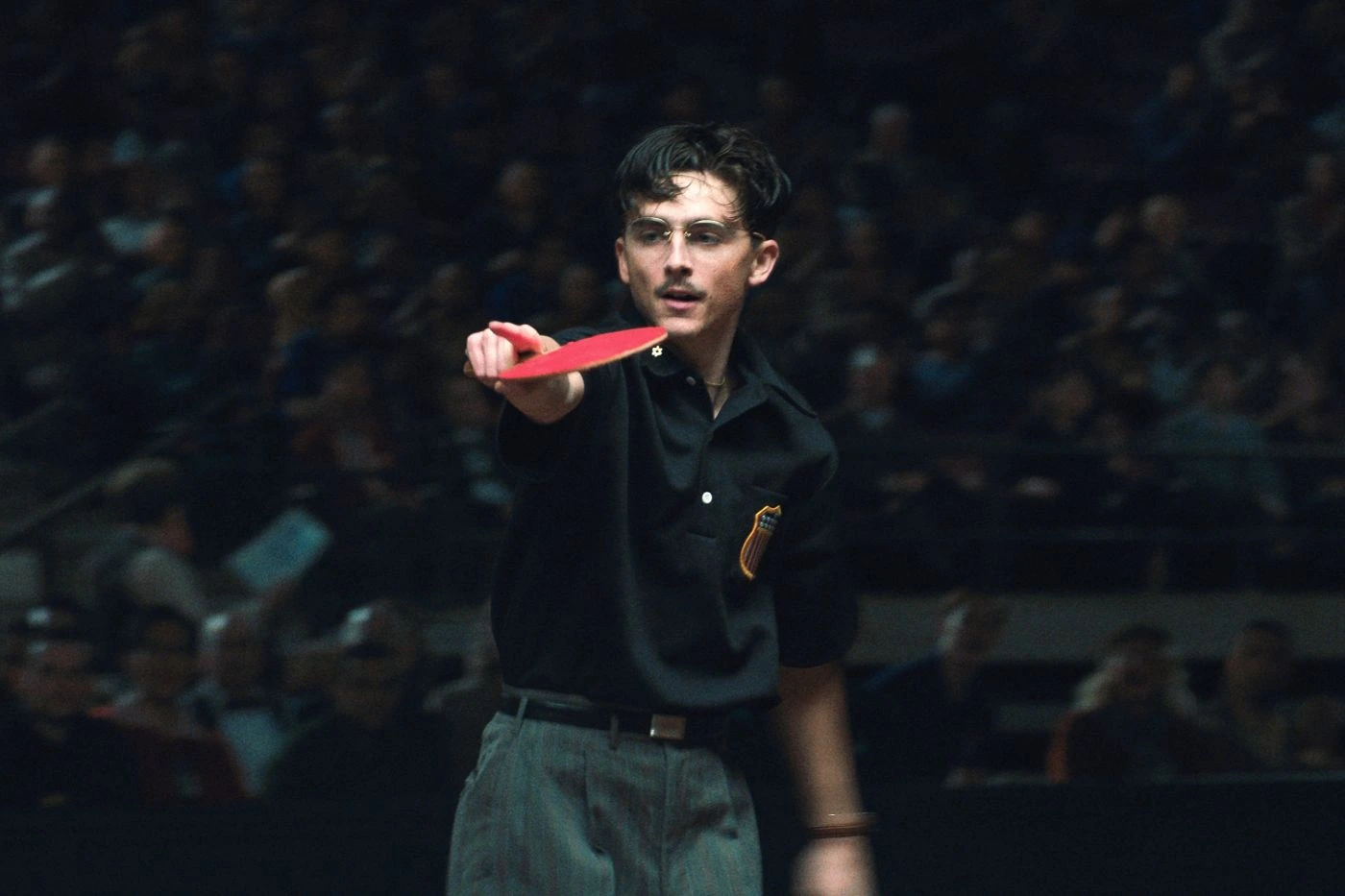 Eine Person in dunkler Kleidung steht in einer Halle vor einem unscharfen Publikum und hält einen roten Tischtennisschläger mit ausgestrecktem Arm nach vorne. Auf dem Hemd ist ein auffälliges Abzeichen zu sehen.