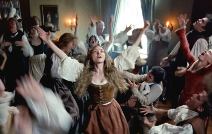 Szenenbild aus "The Testament of Ann Lee": Eine Frau mit langen, gewellten Haaren trägt ein braunes Kleid mit weißer Bluse und steht mit ausgebreiteten Armen im Mittelpunkt eines Raumes. Um sie herum bewegen sich mehrere Personen in historischer Kleidung, viele mit erhobenen Armen. Im Hintergrund sind ein Fenster mit blauen Vorhängen, ein Gemälde an der Wand und zwei brennende Kerzenleuchter zu sehen. Die Szene wirkt dynamisch und lebhaft. [erstellt mit KI]