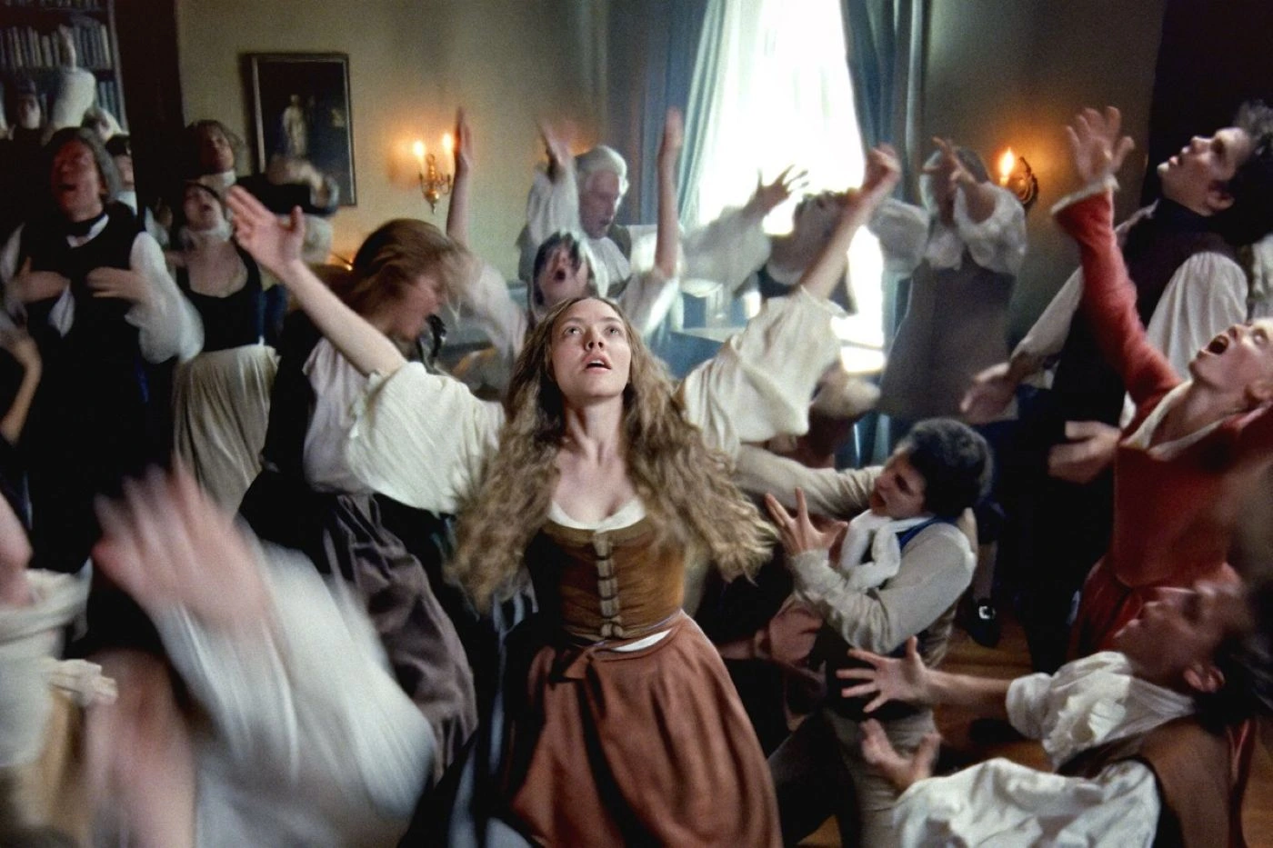 Szenenbild aus "The Testament of Ann Lee": Eine Frau mit langen, gewellten Haaren trägt ein braunes Kleid mit weißer Bluse und steht mit ausgebreiteten Armen im Mittelpunkt eines Raumes. Um sie herum bewegen sich mehrere Personen in historischer Kleidung, viele mit erhobenen Armen. Im Hintergrund sind ein Fenster mit blauen Vorhängen, ein Gemälde an der Wand und zwei brennende Kerzenleuchter zu sehen. Die Szene wirkt dynamisch und lebhaft. [erstellt mit KI]