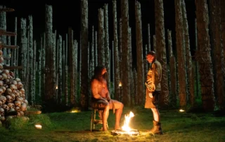 Zwei Personen stehen auf einer Wiese im Schein eines offenen Feuers. Im Hintergrund sind zahlreiche senkrecht aufgestellte Baumstämme zu sehen, die in regelmäßigen Abständen angeordnet sind. Links im Bild befindet sich ein großer Stapel menschlicher Schädel. Die Szene ist nachts und wird von mehreren kleinen Lichtquellen beleuchtet. [erstellt mit KI]
