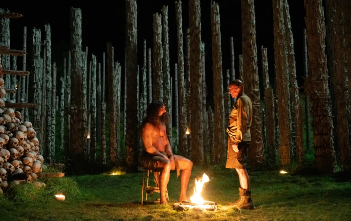 Zwei Personen stehen auf einer Wiese im Schein eines offenen Feuers. Im Hintergrund sind zahlreiche senkrecht aufgestellte Baumstämme zu sehen, die in regelmäßigen Abständen angeordnet sind. Links im Bild befindet sich ein großer Stapel menschlicher Schädel. Die Szene ist nachts und wird von mehreren kleinen Lichtquellen beleuchtet. [erstellt mit KI]