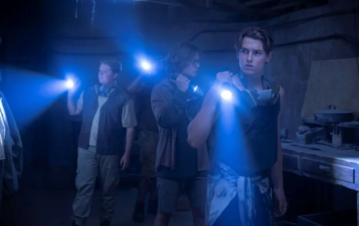 Drei Personen stehen in einem dunklen Raum und halten Taschenlampen, deren Lichtkegel nach vorne gerichtet sind. Die Personen tragen Freizeitkleidung und haben Tücher oder Halstücher um den Hals. Im Hintergrund sind Werkbänke und verschiedene Gegenstände zu erkennen, die auf eine Werkstatt oder einen Kellerraum hindeuten. Das Licht der Taschenlampen erzeugt einen blauen Schein und hebt Staubpartikel in der Luft hervor. [erstellt mit KI]