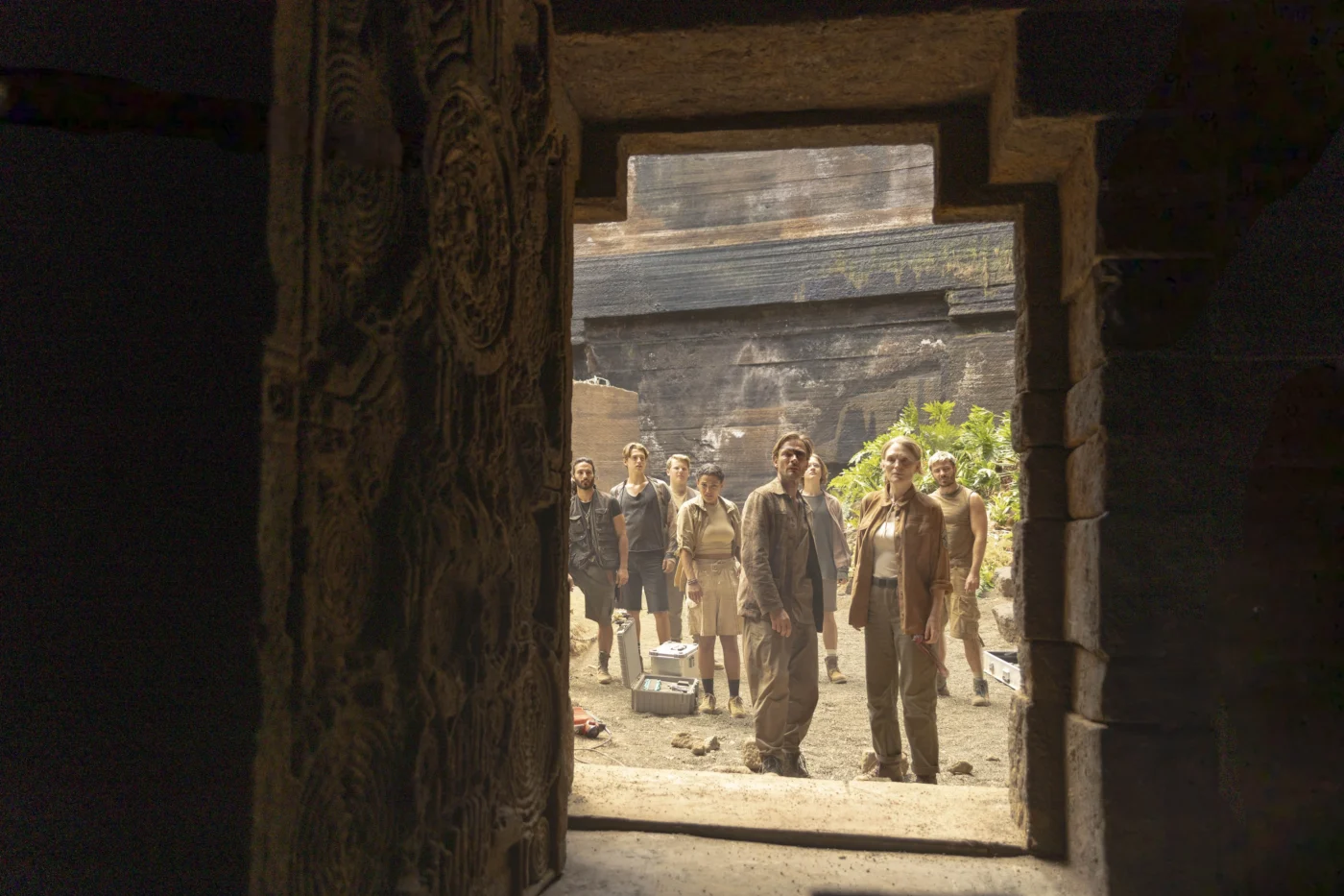 Mehrere Personen stehen in einem offenen, steinernen Durchgang und blicken nach draußen. Die Szene spielt in einer Umgebung mit dunklen Steinwänden und vereinzeltem Grün im Hintergrund. Die Kleidung der Personen ist überwiegend in gedeckten, erdigen Farbtönen gehalten. Die Tür weist reliefartige Muster auf. [erstellt mit KI]