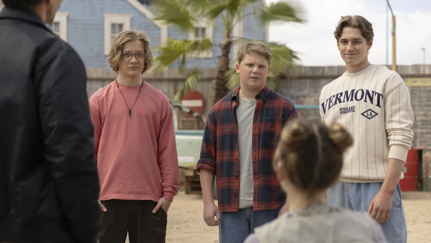 Vier Personen stehen im Freien auf sandigem Boden. Im Hintergrund sind eine blaue Hauswand, eine Palme und ein Verkehrsschild zu sehen. Eine Person trägt ein langärmeliges, cremefarbenes Oberteil mit dunkelblauem Schriftzug und hellblaue Jeans. Eine weitere Person trägt ein kariertes Hemd über einem roten T-Shirt. Die dritte Person trägt ein rosafarbenes Langarmshirt und eine Halskette. Im Vordergrund sitzt eine Person mit zusammengebundenen Haaren und gestreiftem Oberteil mit dem Rücken zur Kamera. [erstellt mit KI]