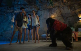 Ein Schimpanse in rotem Oberteil steht aufrecht auf einer Terrasse vor einer Gruppe von Menschen. Im Hintergrund ist eine Felswand beleuchtet, daneben ein Sofa mit Kissen und eine Lampe. Die Szene ist abends oder nachts ausgeleuchtet. [erstellt mit KI]