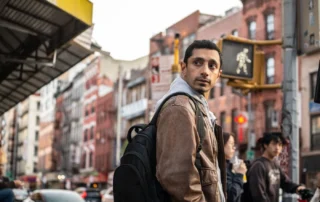 Eine Person mit brauner Jacke und schwarzem Rucksack steht an einer Straßenecke in einer Stadt. Im Hintergrund sind mehrstöckige Gebäude mit roten und weißen Fassaden sowie ein beleuchtetes Fußgängerampel-Symbol zu sehen. Autos und weitere Personen befinden sich auf der Straße. [erstellt mit KI]