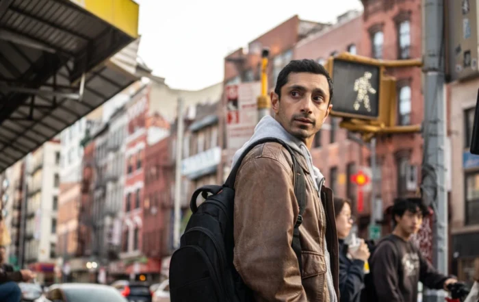 Eine Person mit brauner Jacke und schwarzem Rucksack steht an einer Straßenecke in einer Stadt. Im Hintergrund sind mehrstöckige Gebäude mit roten und weißen Fassaden sowie ein beleuchtetes Fußgängerampel-Symbol zu sehen. Autos und weitere Personen befinden sich auf der Straße. [erstellt mit KI]