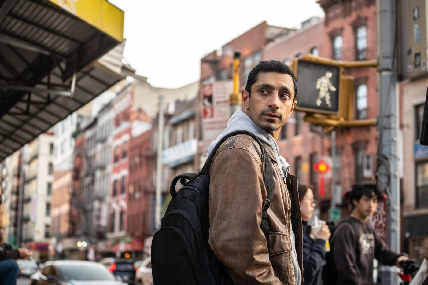 Eine Person mit brauner Jacke und schwarzem Rucksack steht an einer Straßenecke in einer Stadt. Im Hintergrund sind mehrstöckige Gebäude mit roten und weißen Fassaden sowie ein beleuchtetes Fußgängerampel-Symbol zu sehen. Autos und weitere Personen befinden sich auf der Straße. [erstellt mit KI]