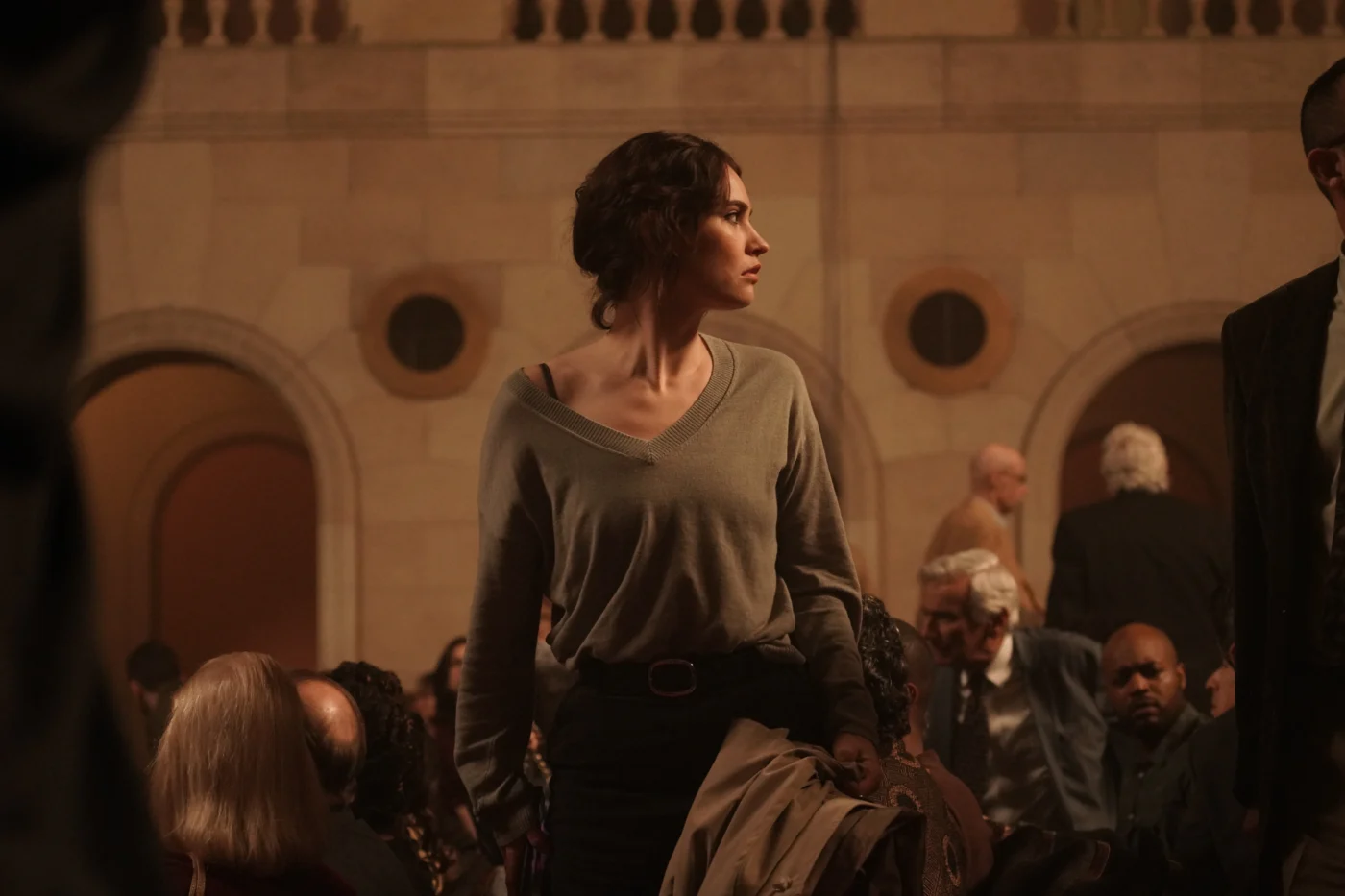 Eine Person mit dunklem, lockigem Haar steht in einem großen Saal mit hohen, steinernen Wänden und Rundbögen. Sie trägt einen grauen Pullover mit V-Ausschnitt und einen dunklen Rock oder eine Hose. Im Hintergrund sind mehrere Menschen zu sehen, einige sitzen, andere stehen. Die Beleuchtung ist warm und gedämpft. [erstellt mit KI]