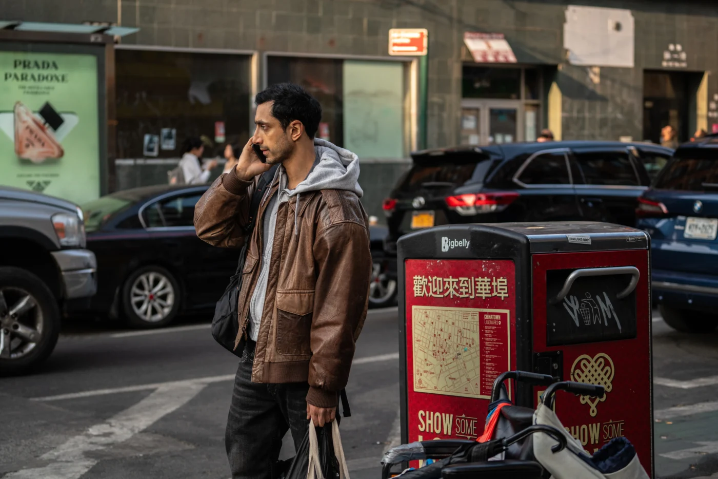 Eine Person steht auf einem Bürgersteig vor einem Mülleimer mit auffälliger roter Werbefläche und chinesischen Schriftzeichen. Die Person trägt eine braune Lederjacke über einem grauen Kapuzenpullover und hält eine Tasche in der linken Hand. Im Hintergrund sind mehrere geparkte Autos und ein Geschäft mit Schaufenster zu sehen. Die Szene spielt sich bei Tageslicht in einer städtischen Umgebung ab. [erstellt mit KI]