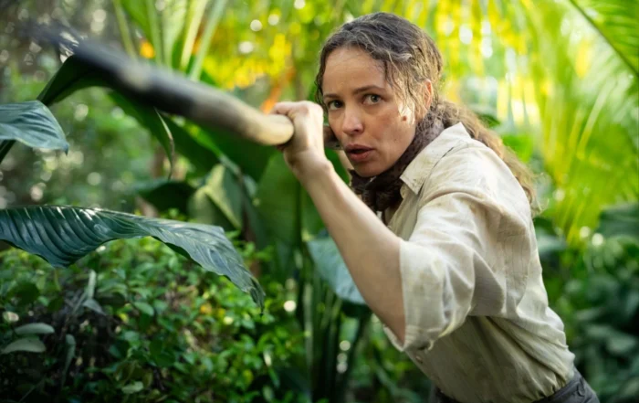 Eine Person mit langen, lockigen Haaren trägt ein helles, langärmliges Hemd und hält einen langen Stock oder Speer. Die Umgebung ist dicht bewachsen mit großen, grünen Blättern und tropischen Pflanzen. Sonnenlicht fällt durch das Blattwerk und erzeugt helle und schattige Bereiche. [erstellt mit KI]