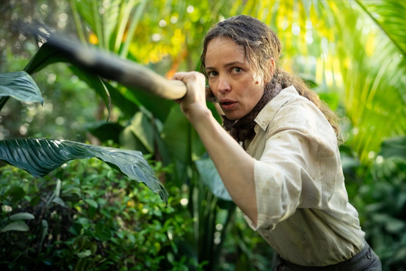 Eine Person mit langen, lockigen Haaren trägt ein helles, langärmliges Hemd und hält einen langen Stock oder Speer. Die Umgebung ist dicht bewachsen mit großen, grünen Blättern und tropischen Pflanzen. Sonnenlicht fällt durch das Blattwerk und erzeugt helle und schattige Bereiche. [erstellt mit KI]