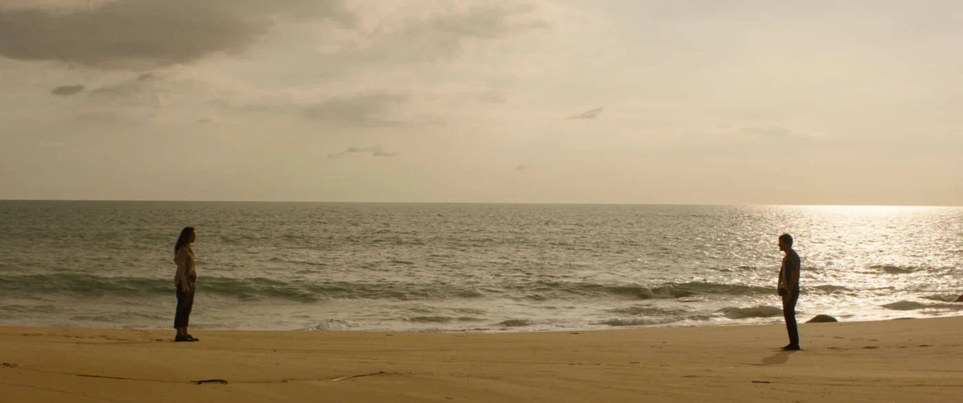Zwei Personen stehen mit großem Abstand zueinander am Rand eines weitläufigen Sandstrandes. Der Himmel ist bewölkt, das Licht wirkt gedämpft. Im Hintergrund erstreckt sich das ruhige Meer bis zum Horizont. [erstellt mit KI]