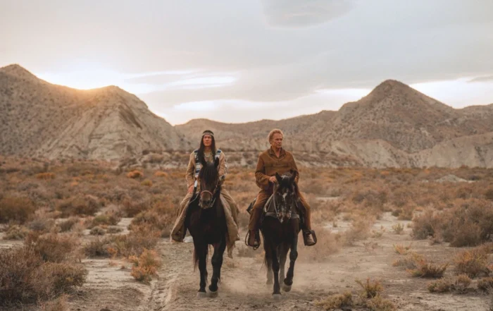 Zwei Personen reiten nebeneinander auf dunklen Pferden durch eine trockene, steinige Landschaft mit niedrigen Büschen. Im Hintergrund sind sanft geschwungene Hügel und ein bewölkter Himmel zu sehen. Die Kleidung wirkt robust und ist in gedeckten Farben gehalten. Staub wird von den Hufen der Pferde aufgewirbelt. [erstellt mit KI]