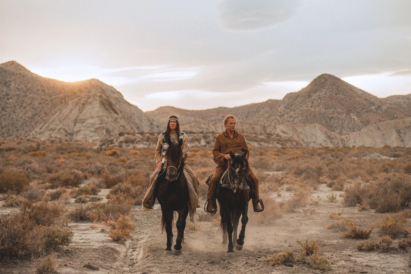 Zwei Personen reiten nebeneinander auf dunklen Pferden durch eine trockene, steinige Landschaft mit niedrigen Büschen. Im Hintergrund sind sanft geschwungene Hügel und ein bewölkter Himmel zu sehen. Die Kleidung wirkt robust und ist in gedeckten Farben gehalten. Staub wird von den Hufen der Pferde aufgewirbelt. [erstellt mit KI]