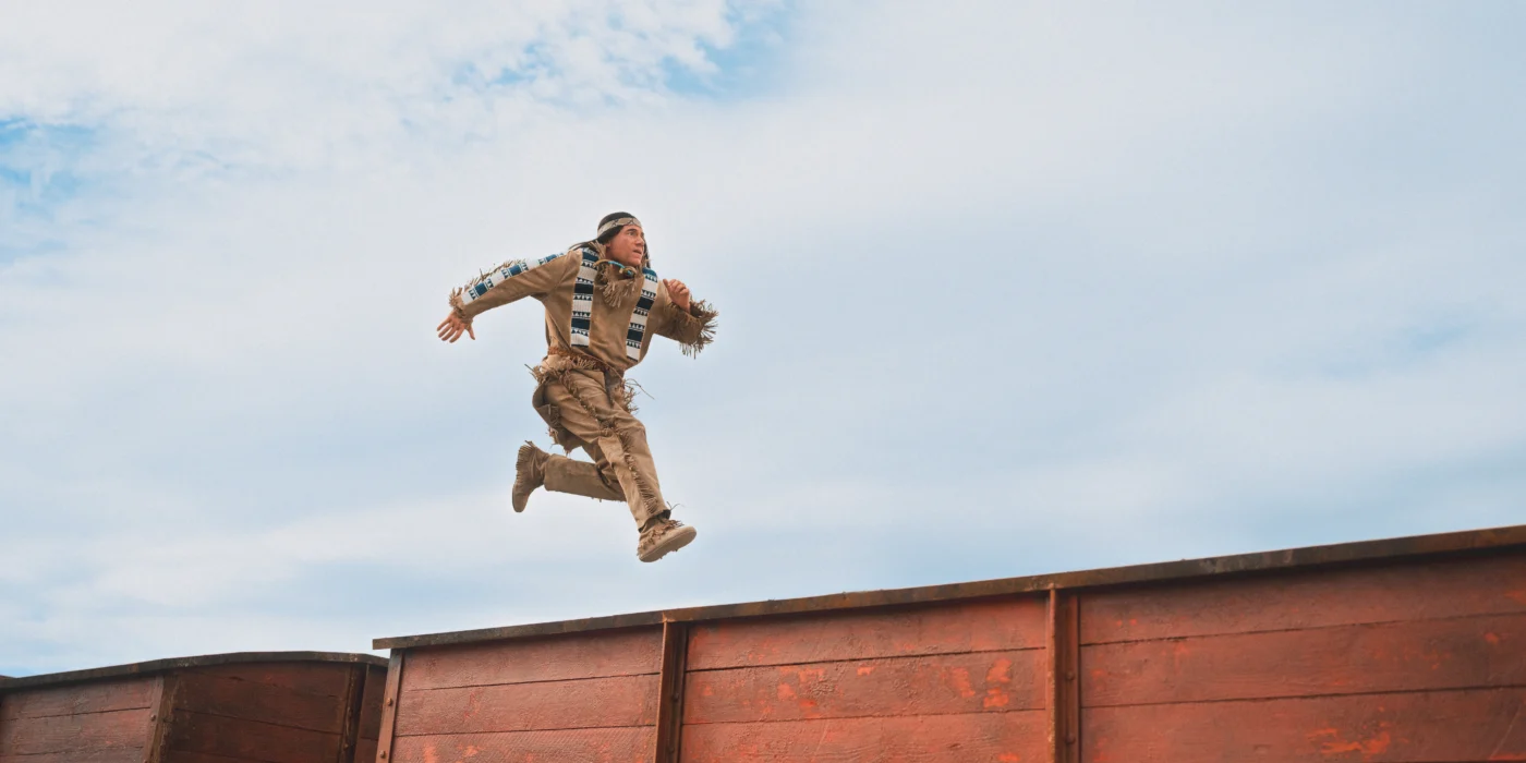Eine Person in beigefarbener Kleidung springt mit ausgestreckten Armen und Beinen über einen braunen Holzzaun. Der Himmel ist leicht bewölkt und bildet den Hintergrund. [erstellt mit KI]