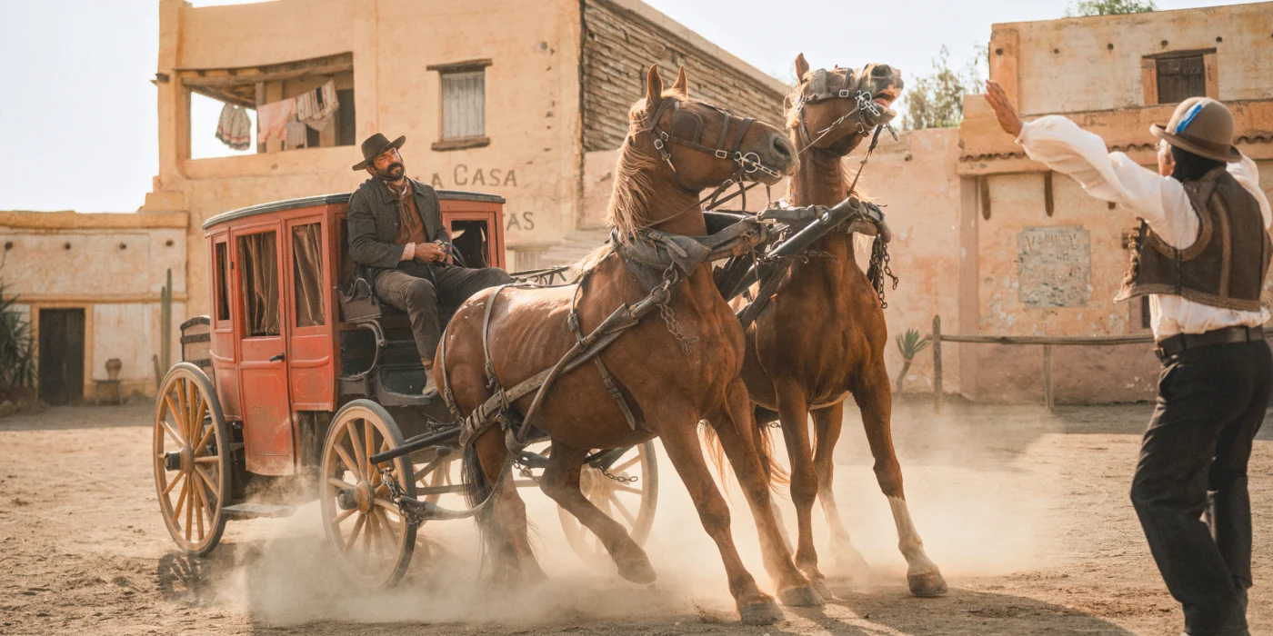 Eine Postkutsche mit zwei braunen Pferden steht auf einem staubigen Platz vor einem Gebäude mit der Aufschrift 'LA CASA'. Der Kutscher trägt einen dunklen Hut und hält die Zügel, während die Pferde aufgeregt und angespannt wirken. Ein Mann mit Hut und Weste steht vor der Kutsche und hebt beide Arme, als wolle er die Pferde beruhigen oder anhalten. Im Hintergrund sind weitere Gebäude im Stil eines Western-Dorfes zu sehen. [erstellt mit KI]