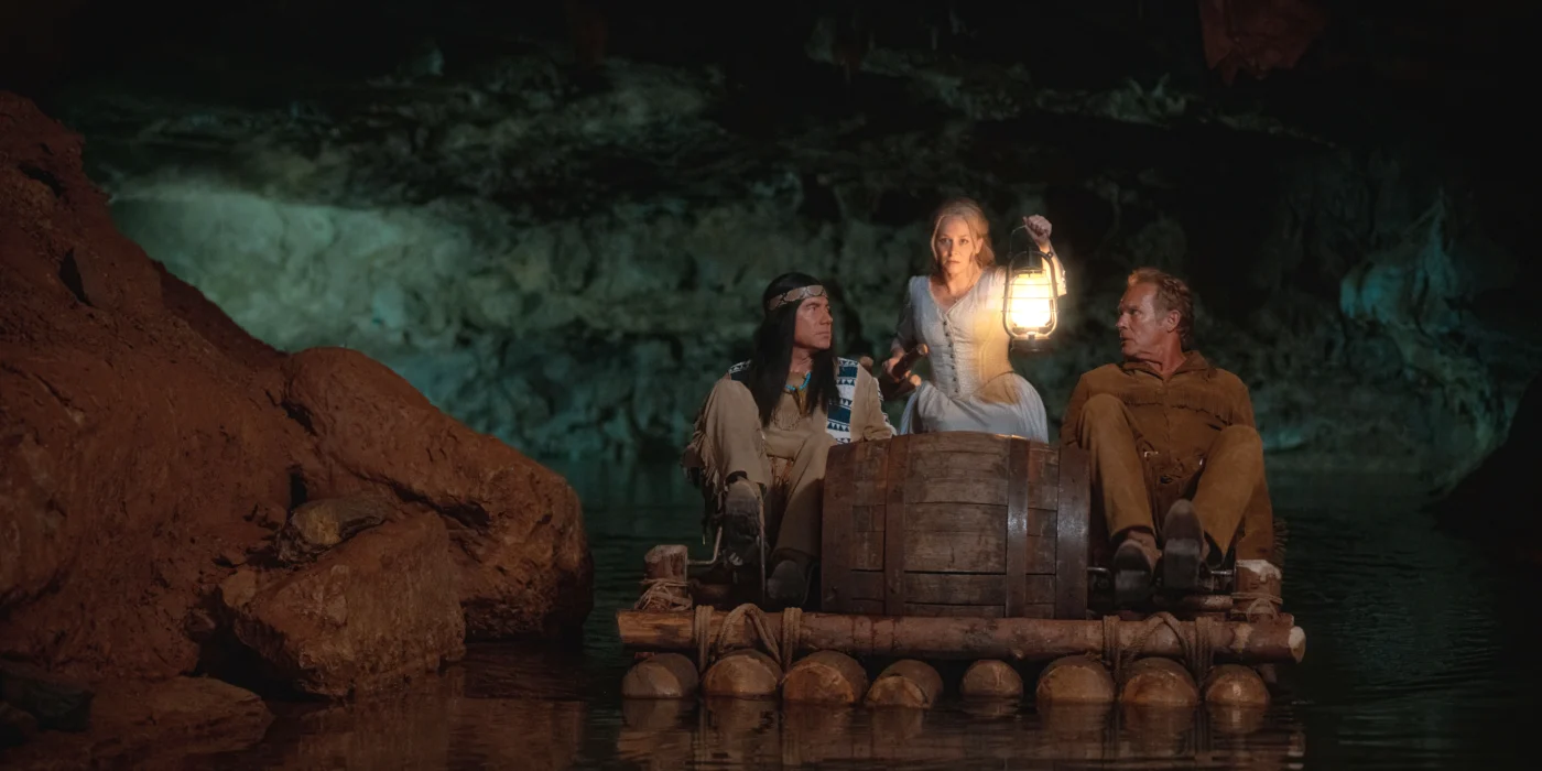 Drei Personen sitzen auf einem Floß aus Holz und Fässern in einer Höhle. Die mittlere Person hält eine leuchtende Laterne hoch. Die Umgebung ist von Felsen und Wasser geprägt, im Hintergrund ist eine beleuchtete Höhlenwand zu sehen. [erstellt mit KI]