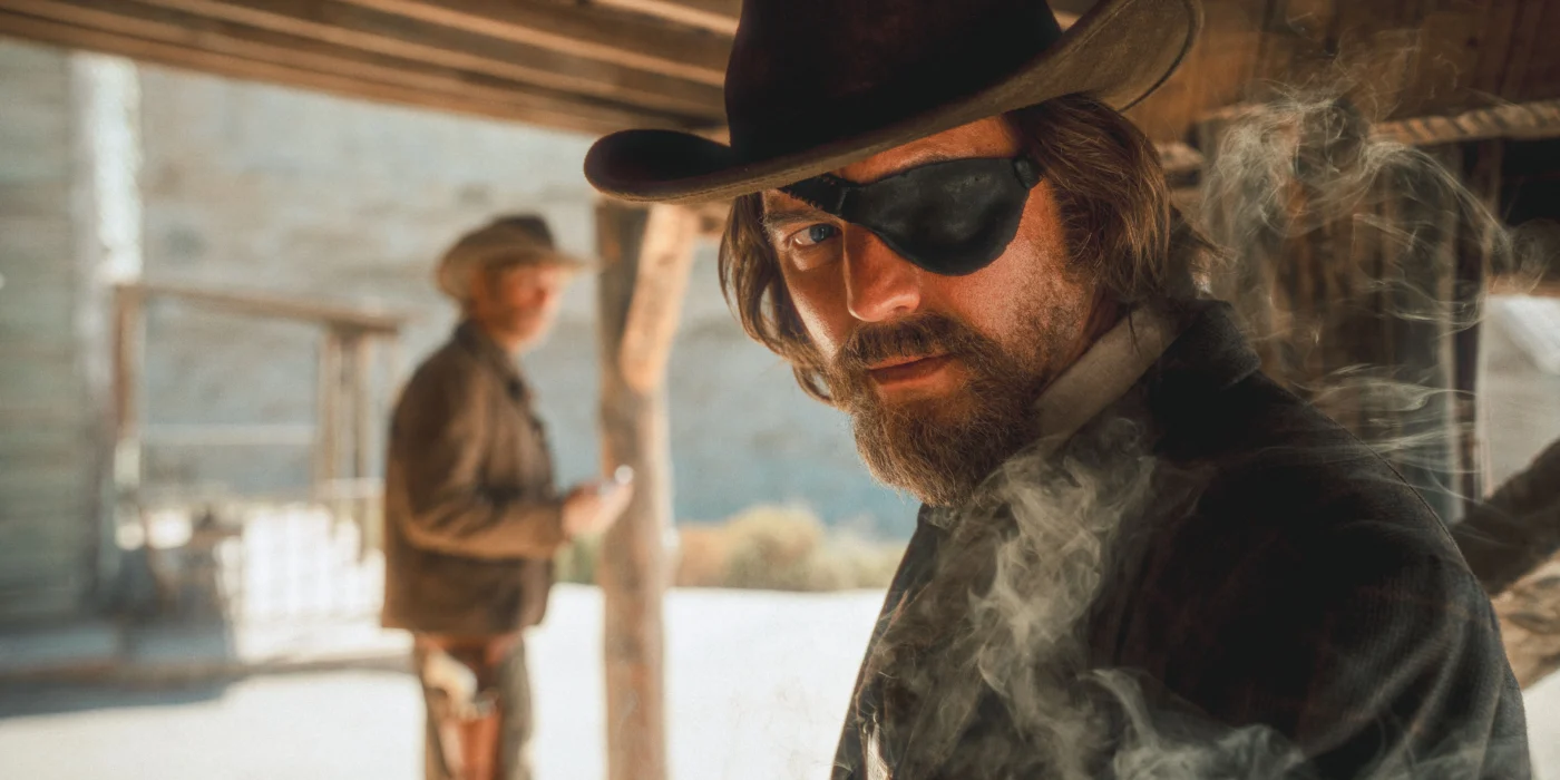 Zwei Personen stehen unter einem hölzernen Vordach in einer Westernkulisse. Beide tragen Cowboyhüte und typische Westernkleidung. Im Vordergrund sind Rauchschwaden sichtbar, die die Szene atmosphärisch wirken lassen. Die Beleuchtung ist warm und betont die rustikale Umgebung mit Holzbalken und Steinmauer im Hintergrund. [erstellt mit KI]