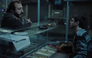 Zwei Personen befinden sich an einer Theke in einem Imbiss oder Restaurant. Eine Person sitzt auf der rechten Seite und trägt einen gestreiften Pullover. Die andere Person lehnt auf der linken Seite an der Theke und hält eine Zigarette in der Hand. Auf der Theke liegen ein Stapel Papiere und ein rundes Tablett. Im Vordergrund sind mehrere Edelstahlschalen mit Speisen in einer Auslage zu sehen. Die Szene ist schwach beleuchtet und wirkt kühl. [erstellt mit KI]