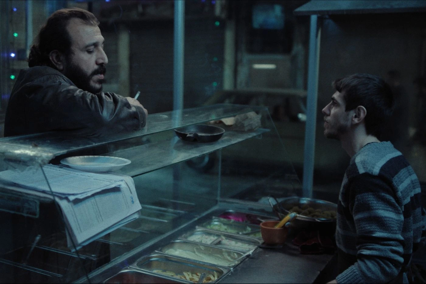 Zwei Personen befinden sich an einer Theke in einem Imbiss oder Restaurant. Eine Person sitzt auf der rechten Seite und trägt einen gestreiften Pullover. Die andere Person lehnt auf der linken Seite an der Theke und hält eine Zigarette in der Hand. Auf der Theke liegen ein Stapel Papiere und ein rundes Tablett. Im Vordergrund sind mehrere Edelstahlschalen mit Speisen in einer Auslage zu sehen. Die Szene ist schwach beleuchtet und wirkt kühl. [erstellt mit KI]